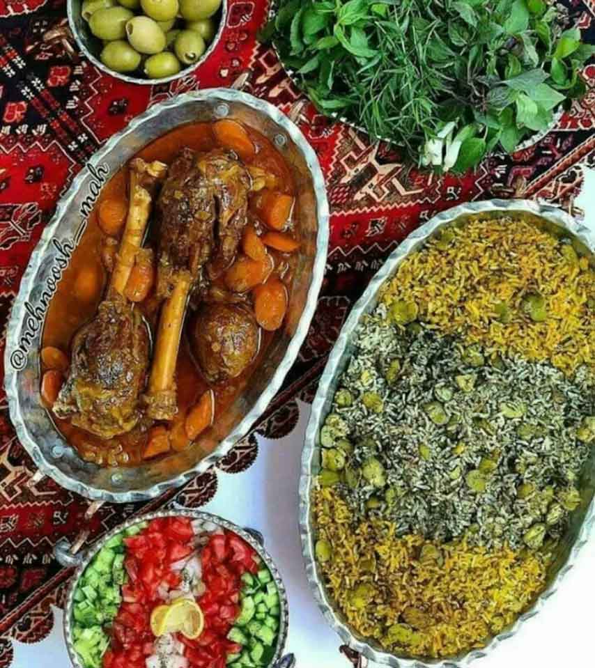 A plate of Mahiche (braised lamb shank) with rice, tea, and fresh herbs on a patterned tablecloth.